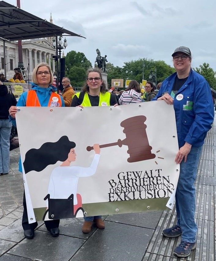Julia Moser, Paulina Spelbrink und Heidemarie Egger, 
        halten eine 150x100 cm große Plane, auf der die Illustration gedruckt wurde. Im Hintergrund ist das Parlament, eine 
        Menschenansammlung und eine Bühne. Auf dieser wurde die UN-BRK durch Vertreter*innen verschiedener Organisationen Artikel 
        für Artikel vorgelesen.Beschreibung der Illustration: Eine Frau mit wehenden schwarzen Haaren hält einen überdimensionierten 
        Richterhammer in ihrer rechten Hand. Diese werden zum Beispiel in den USA verwendet, sind aber vor allem durch Filme und Serien 
        auch bei uns bekannt. Er steht hier symbolisch für die Menschenrechte. Man sieht die Frau von hinten (leicht schräg allerdings,  
        damit es dynamischer aussieht), sie blickt zur Seite Richtung Hammer. Ihr Arm ist dabei kampfbereit erhoben, allerdings, ohne 
        aggressiv oder bedrohlich zu wirken. Das wird auch durch ihren Gesichtsausdruck unterstrichen. Insgesamt wirkt ihre Körperhaltung 
        entschlossen und sie strahlt Stärke aus. Sie nutzt einen vereinfacht gezeichneten schwarzen Rollstuhl. Dieser ist im Verhältnis 
        zur Person eher klein. Das liegt vor allem an der kurzen Rückenlehne. Aber auch daran, dass das Bild unterhalb der Sitzfläche 
        endet und so von den Rädern nur der obere Teil zu sehen ist. Es war mir wichtig, dass der Rollstuhl nicht der Fokuspunkt des 
        Bildes ist. Denn Rollstuhlnutzer*innen machen öfter die Erfahrung, dass in der Interaktion mit anderen, nur noch der Rollstuhl 
        gesehen wird und nicht die Person. Der Rollstuhl wird dafür nicht versteckt, sondern er ist als selbstverständlicher Teil der 
        Lebensrealität der Person einfach da, ohne von dieser abzulenken oder extra thematisiert zu werden. Es befindet sich auch ein Text 
        im Bild. Durch die Anordnung unterhalb des Hammers wird angedeutet, dass die Frau im Rollstuhl im Vordergrund und der Text ein 
        kleines Stück entfernt ist. Sie bewegt sich also darauf zu. Es steht Gewalt, Barrieren, Diskriminierung und Exklusion. Die Wörter 
        sind übereinander angeordnet, eins steht auf dem anderen. Man sieht, dass sie schon mit dem Hammer bearbeitet worden sind. Sie 
        haben Sprünge, Stücke sind abgebrochen und manche Buchstaben sind auch schon umgekippt. Das Bild stellt also den Kampf gegen 
        Ausgrenzung von Menschen mit Behinderungen mithilfe der UN-Behindertenrechtskonvention dar.Der Stil der Illustration ist 
        zweidimensional. Die umrandenden Linien sind klar und sind farblich an die Flächen angepasst. Der Hintergrund ist in zwei Teile 
        geteilt. Er ist hauptsächlich hellgelb mit etwas olivgrün im unteren Bereich. Richterhammer sind traditionell aus Holz, weshalb 
        er braun dargestellt ist. Die Frau trägt ein weißes Shirt und eine korallfarbene Hose. Die Farben wirken neutraler, aber 
        gleichzeitig hoffnungsfroh. Wie schon bei der Beschreibung der Frau angesprochen, war meine Intention zwar eine gewisse 
        Kampfbereitschaft, aber nicht im Sinne eines Aufstandes oder ähnlichem, sondern im Sinne einer fairen und gerechten 
        Rechtssprechung basierend auf klar formulierten Menschenrechten.