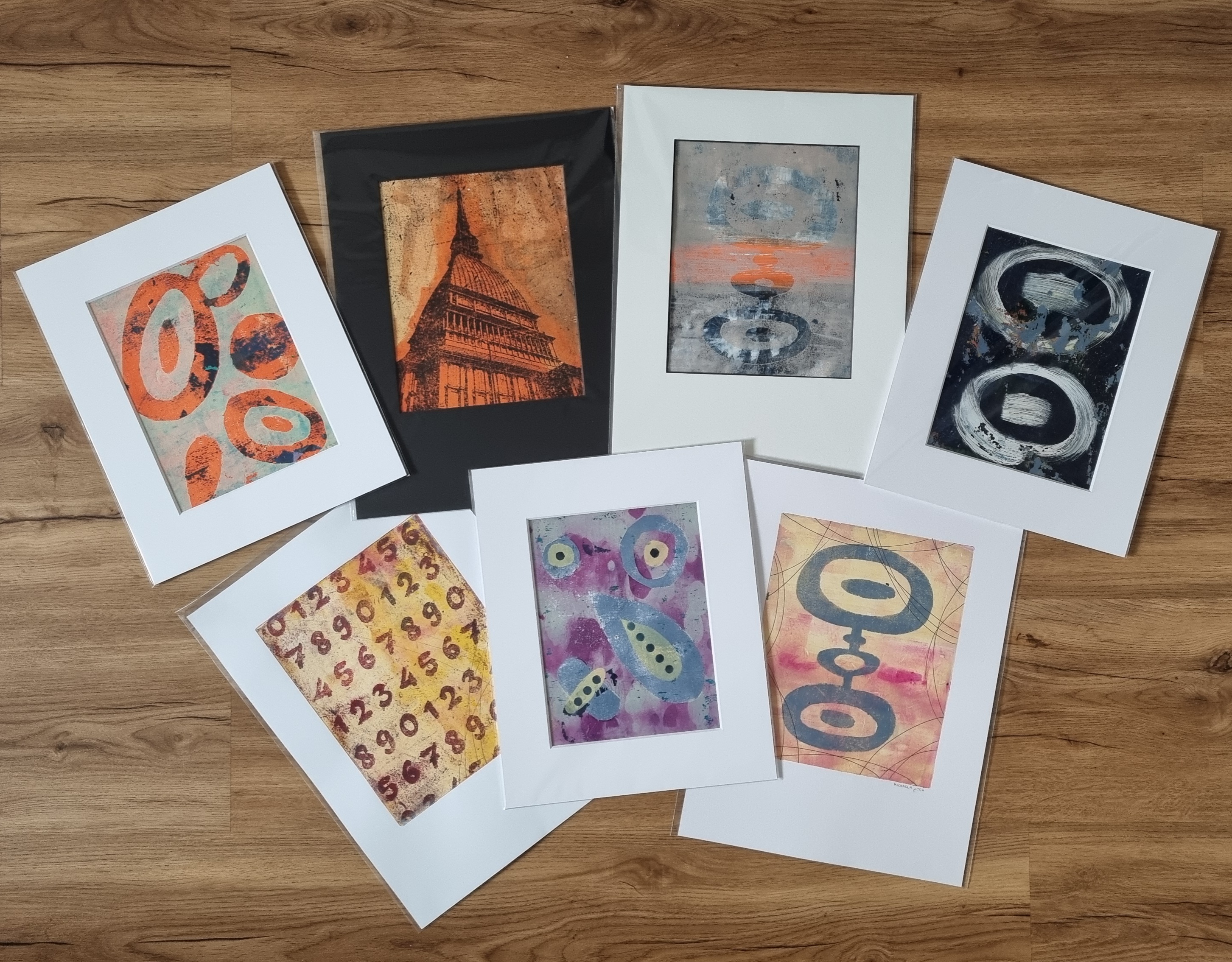 Kurzbeschreibung: Sieben verschiedene Gelplattendrucke liegen schön aufgelegt auf einem hölzernen Untergrund. Sie sind in Schutzfolien eingepackt und manche haben ein Passepartout.
            Langbeschreibung: Bild 1: runde und ovale Formen in Orange mit schwarzen Artefakten sind über die ganze Fläche verteilt. Der Hintergrund ist neapelgelbgrün mit orangenen Untertönen. Bild 2: ein Fototransfer von einem Gebäude in Turin. Die Linien sind schwarz, der Hintergrund orange und gelb. Bild 3: eine Form die aus 4 ovalen Formen zusammengefügt ist. Die oberste und unterste Form sind am größten, die zweite ist am kleinsten. Die Farben haben dem Bild den Titel gegeben Sonnenaufgang am Wasser gegeben. Es sieht aus wie ein Himmel mit Wolken, der sich im Wasser spiegelt, während am Horizont die Sonne in orange-gelb aufgeht. Bild 4: Dieses Bild ist nicht mit Masken gemacht, sondern mit dem Pinsel. Die dominante Form sind zwei Ovale, jeweils mit einem Strich in der Mitte. Die Pinselstriche sind weiß, der Hintergrund dunkelblau. Das Bild ist mit Farbresten vom vorherigen Druck, die auf der Platte geblieben sind, übersäht. Diese sind in einem graublau, das stellenweise mit orange vermischt ist. Bild 5: Hier sind die Zahlen 0-9 in sich wiederholenden Reihen in rot auf die Platte übertragen worden. Der Hintergrund setzt sich aus unterschiedlichen Gelbtönen zusammen. Insgesamt wirkt es als wäre es älter. Bild 7: Das Bild entstand ebenfalls mit Masken. Die Formen sind in graublau und es entsteht der Eindruck eines Gesichts. Vor allem deshalb, weil die Formen mit kleineren Formen in gelb und dunkelblau übermalt sind. Der Hintergrund ist Magenta mit Beige in unterschiedlichen Schattierungen. Diese entstehen, wenn man Farbkleckse auf die Platte gibt und dann mit der Rolle hoch und runterrollt. Es sieht ein bisschen aus wie Tropfen. Bild 7: die gleiche Form wie Bild 3. Der Hintergrund ist hier gelb und magenta, diesmal von links nach rechts gerollt und die Form is graublau.