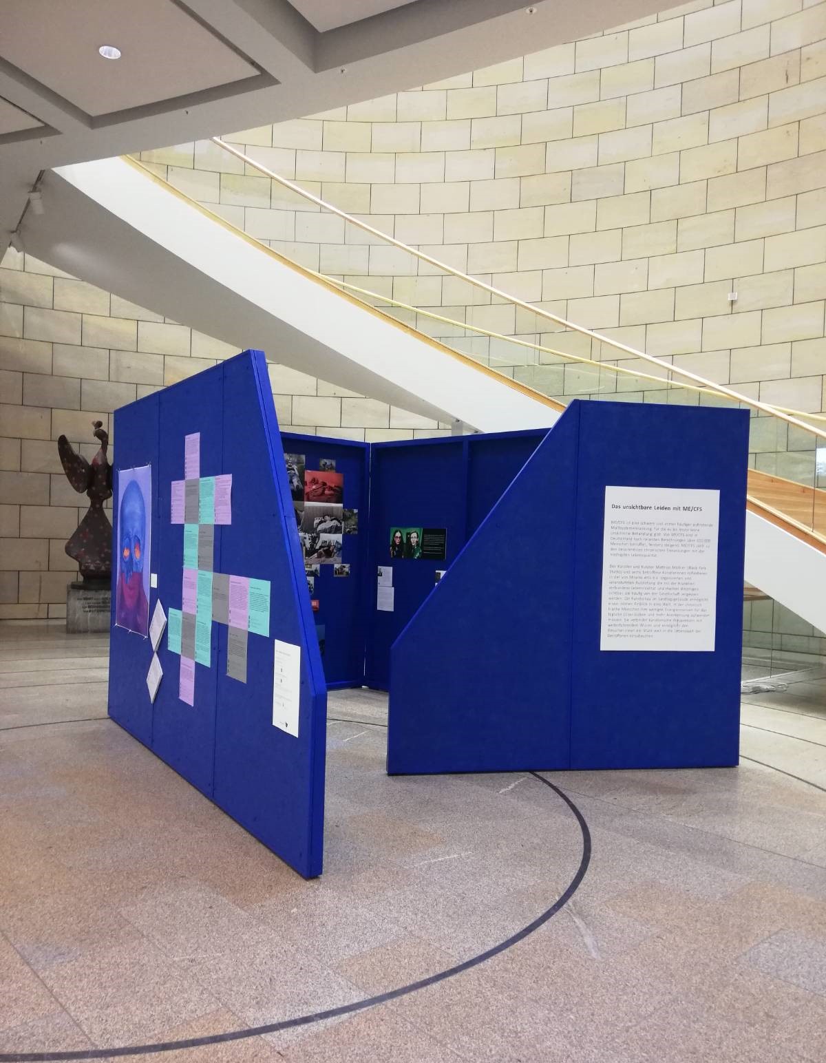 Eine Installation aus blauen Wänden, an der verschiedene Bilder befestigt sind. Im Hintergrund sieht man den Eingangsbereich des Landtagsgebäudes.