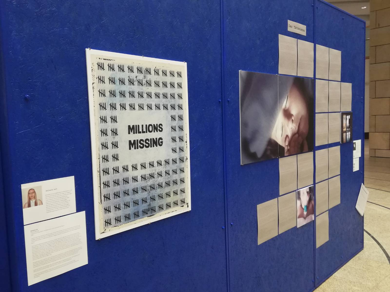 Eine schräg-fotografierte Ansicht von einer der Wände. Vorne ist ein vergrößerter Plakat-Druck eines Millions-Missing-Bildes von mir. Weiter hinten sieht man zwei Fotos von Menschen. Eine hat ein Tuch über den Augen und liegt, die andere bekommt Sauerstoff. Darum verteilt sind außerdem Texte unter dem Titel Der Tätowierer.