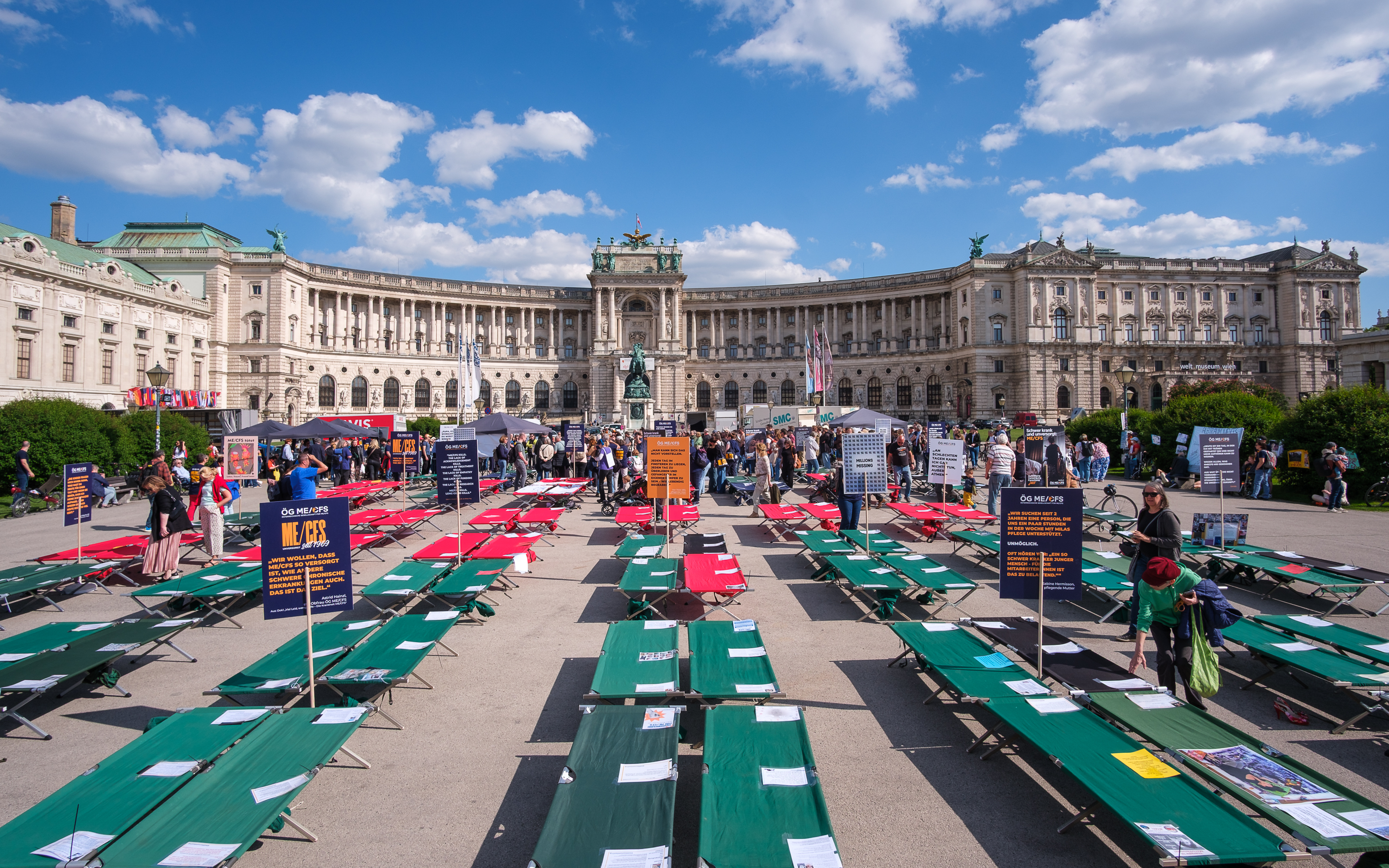 Im Hintergrund ist groß die Hofburg zu sehen, davor stehen 
      viele Feldbetten. Zwischen den Feldbetten sind Menschen, aber auch Plakate aufgestellt. Auf den Feldbetten 
      liegen Botschaften von Menschen, die mit ME/CFS leben.