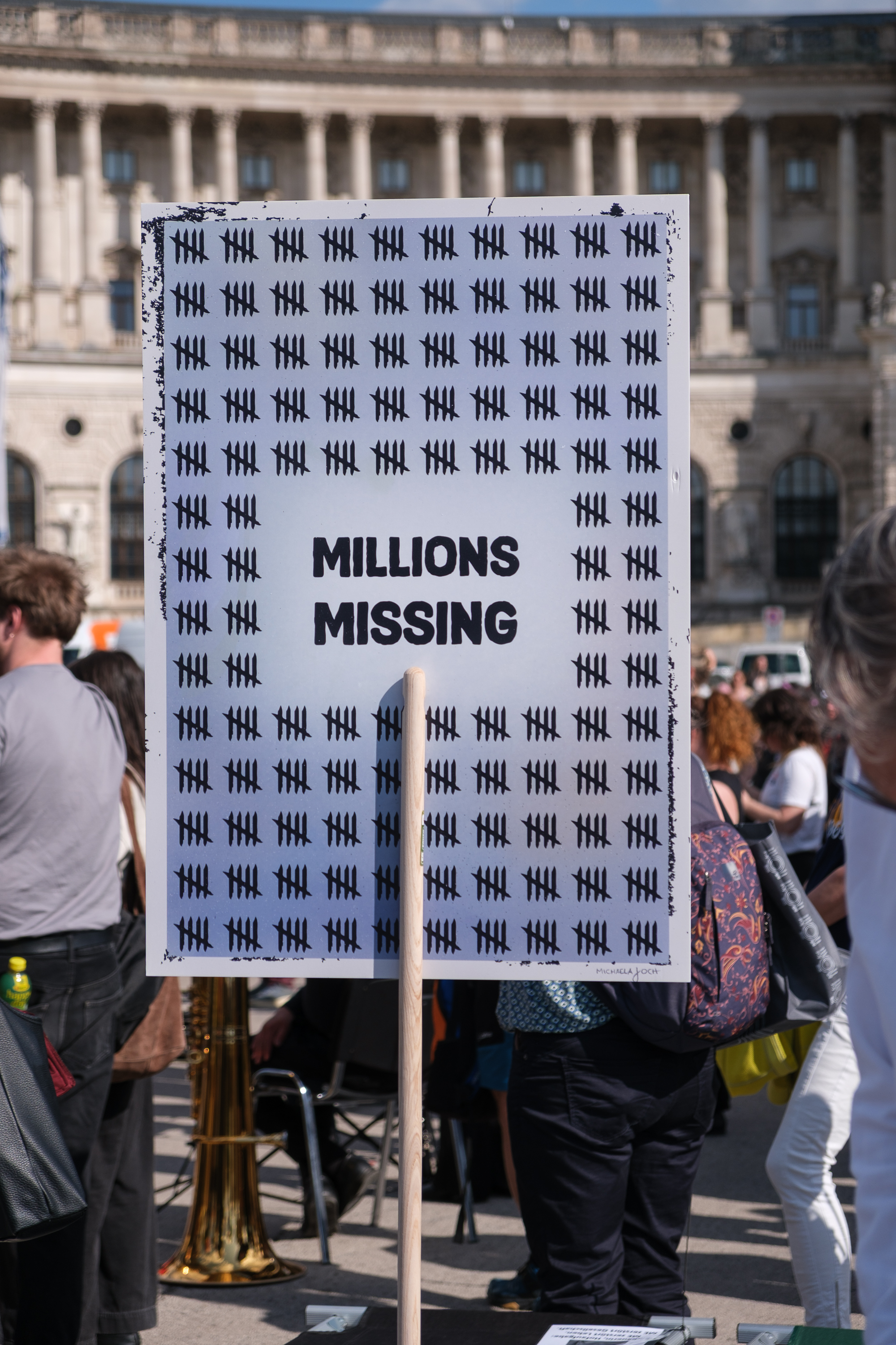 Mein Bild als A1-Plakat gedruckt und auf einem Holzstiel montiert. Es 
     steht zwischen den Feldbetten. Man sieht Personen im Hintergrund. Beschreibung des Plakates: In der Mitte des Bildes steht groß in schwarz 'Millions Missing'. 
     „Millions Missing“ bezieht sich auf all die Menschen, die mit ME/CFS leben und in der Gesellschaft nicht sichtbar sind. Um die in der Gesellschaft 'fehlenden' Menschen 
     darzustellen, ist der Text umgeben von einem ebenfalls schwarzem, universellem Symbol, das sich regelmäßig wiederholt. Das Symbol besteht aus 4 vertikalen Strichen und 
     einem diagonalen Strich, der über die vier verläuft. Dies ist eine andere Art, die Zahl 5 darzustellen. In Summe sind also hunderte Menschen auf dem Bild mit diesen 
     Strichen repräsentiert. Die Farben sind hauptsächlich blau mit etwas orange, grün und gelb. Es wirkt harmonisch und eher positiv.  Aber es hat Elemente, die diesen Eindruck 
     abmildern. Gerade zum Rand hin gibt es schwarze 'Störelemente'.